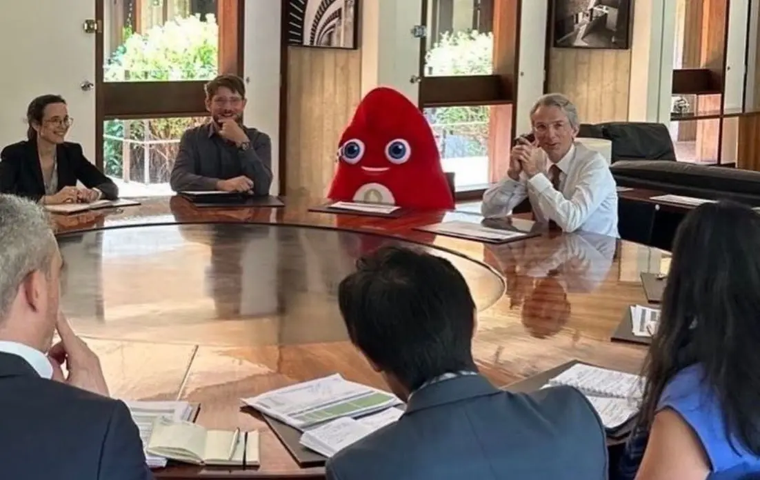 A red, teardrop-shaped cartoon character with large blue eyes and a smiling mouth sits at a large round conference table…