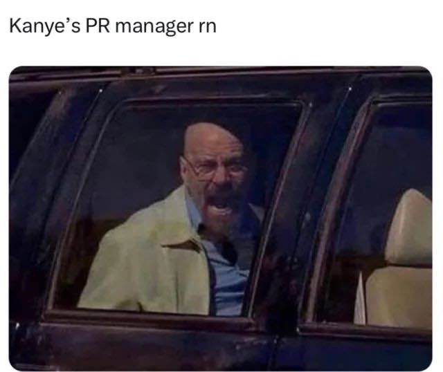 A man with a bald head and glasses, wearing a light-colored jacket over a blue shirt, is leaning out of a car window with…