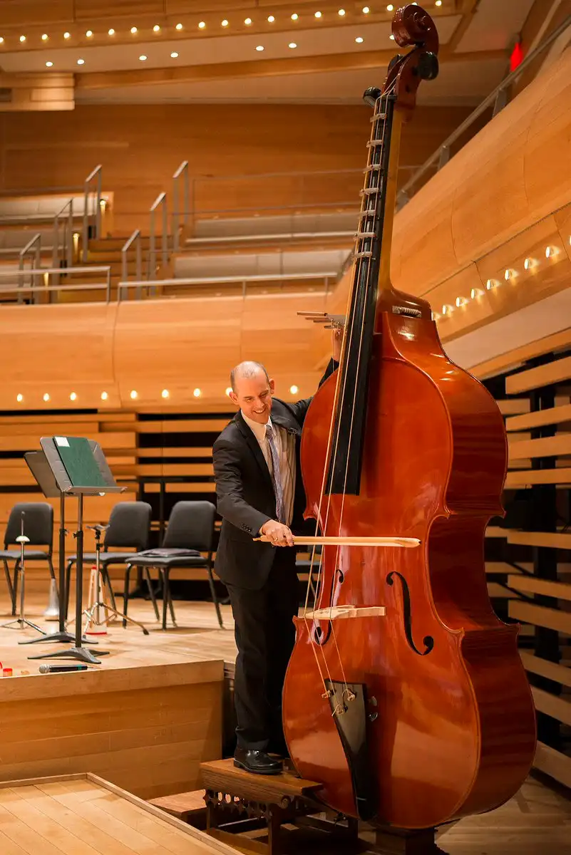A man in a suit stands on a small wooden platform in a concert hall, smiling as he holds a giant upright bass with a bow…