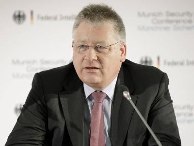A man in a dark suit and glasses, seated at a podium with a microphone, appears to be speaking at a formal event. Behind him…