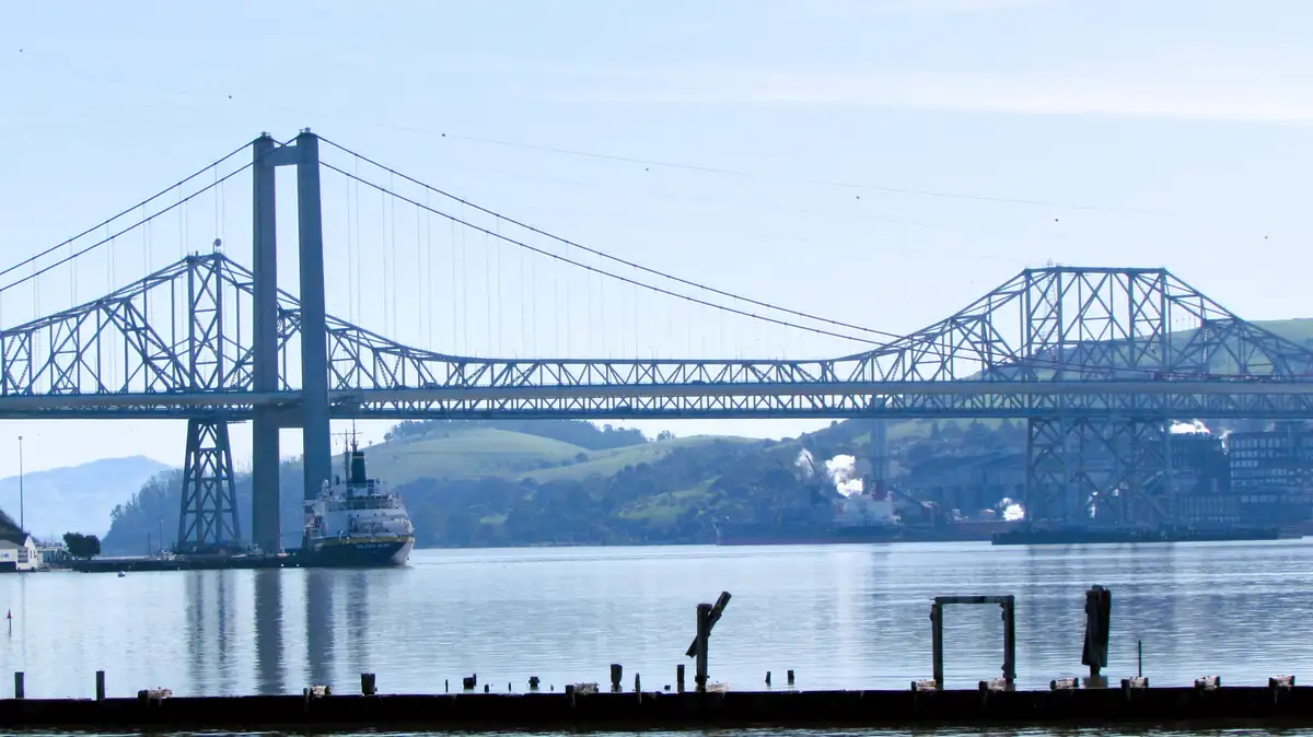 A wide shot of a large suspension bridge spanning a body of water with hills in the background. A large ship named 'GOLDEN…