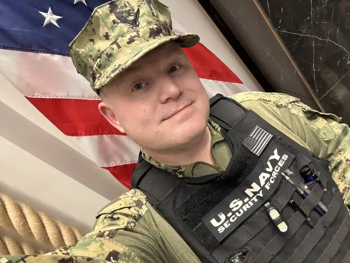 A man in U.S. Navy Security Forces uniform and camouflage cap poses for a selfie with a slight smirk, standing in front of…