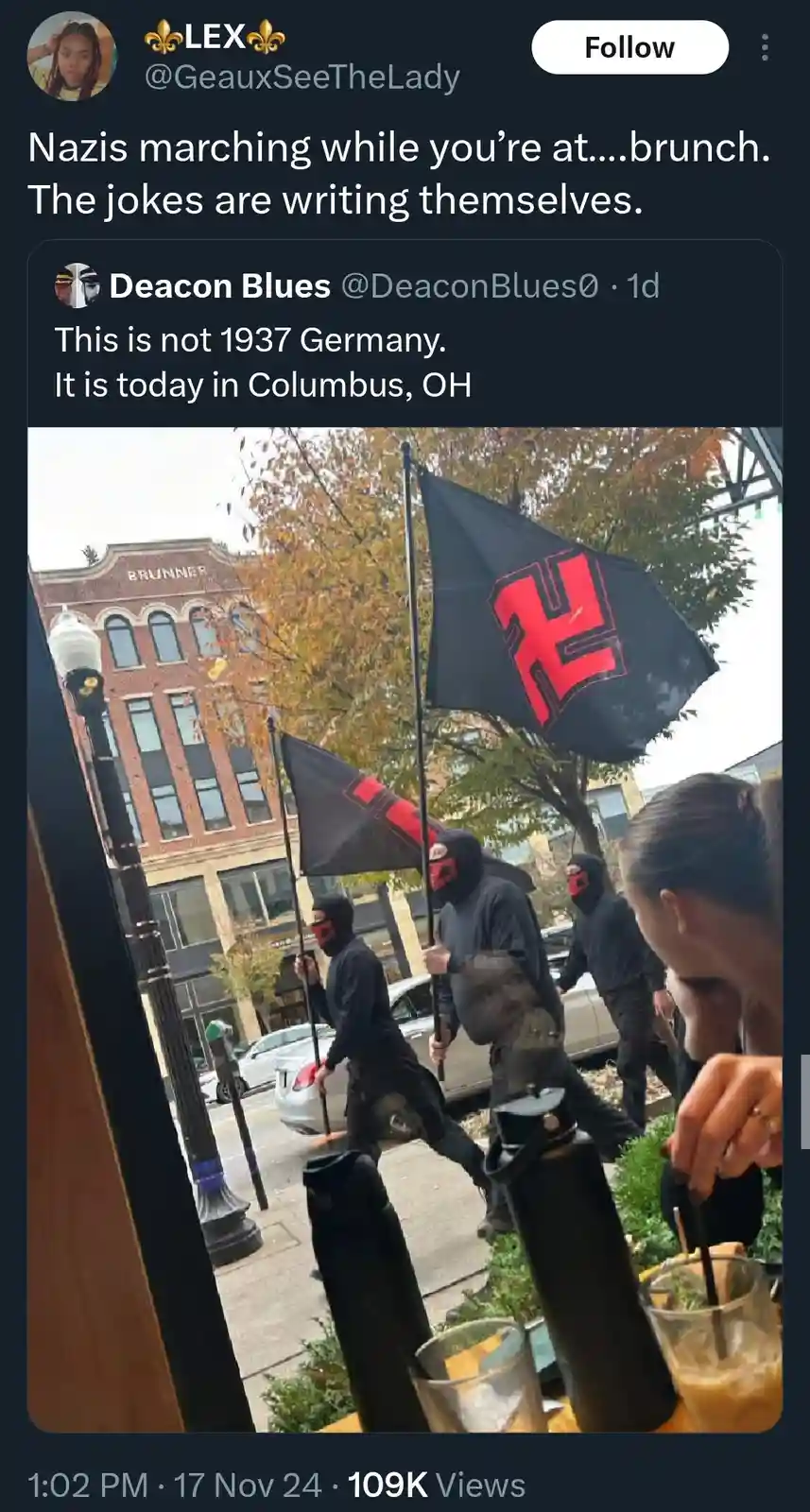 A photo taken from inside a cafe shows several masked individuals marching down a street while holding black flags with red…