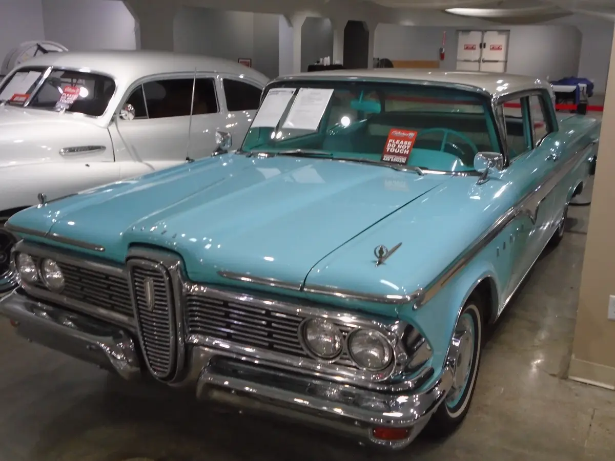 A light blue vintage car with chrome details is displayed indoors, likely in a museum or showroom, with a white classic car…