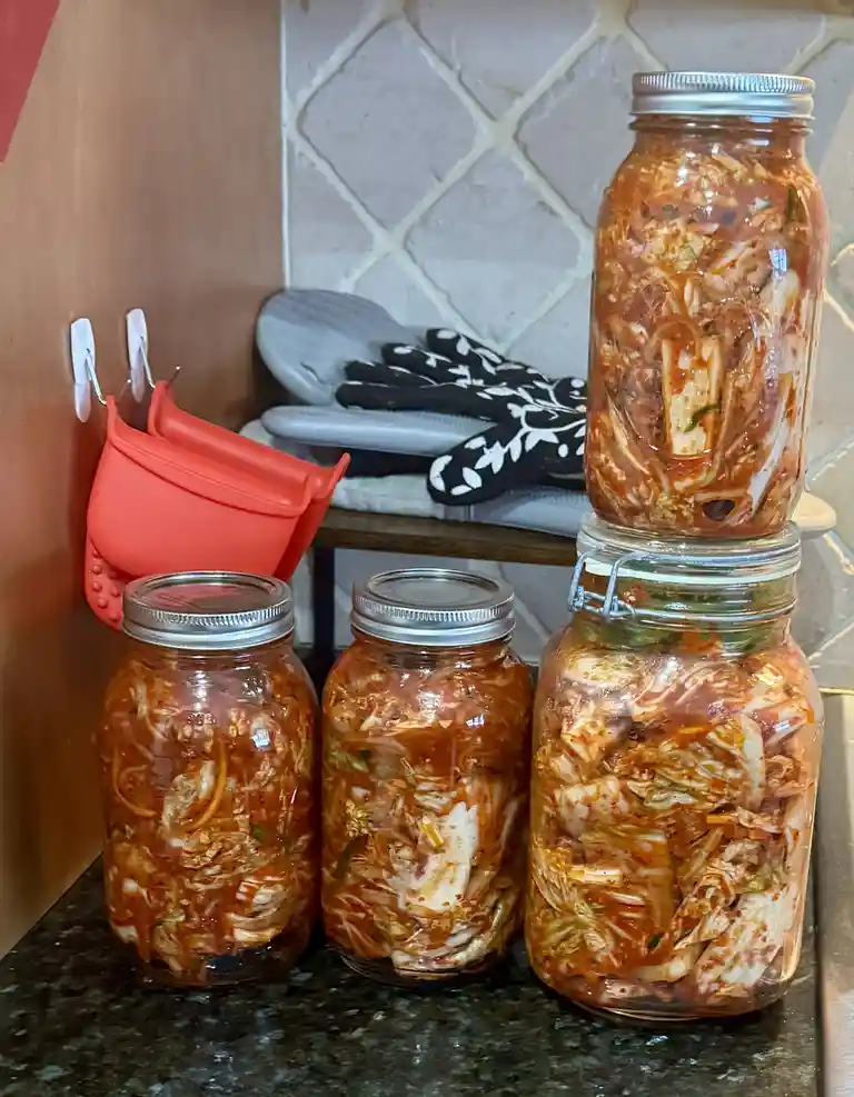 Four glass jars filled with red kimchi sit on a dark countertop, with one jar stacked atop another. In the background, a…