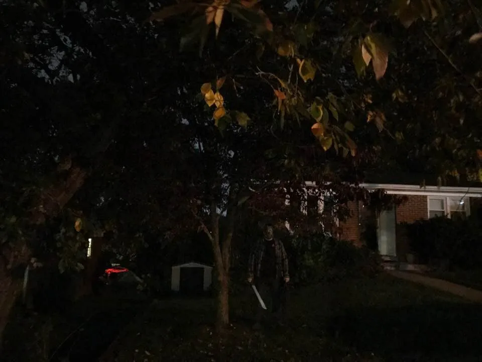 A person wearing a dark mask and holding a large knife stands in a dimly lit yard at night, partially obscured by trees. A…