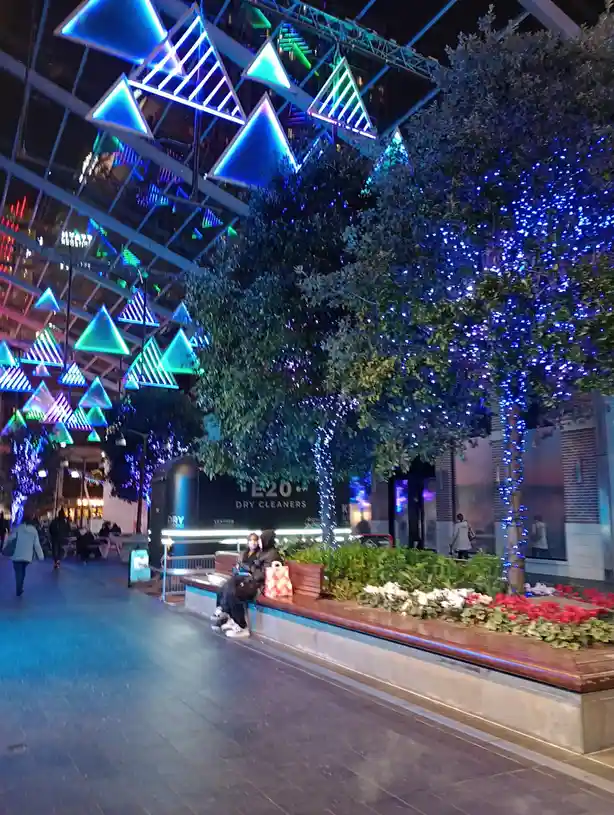 A nighttime photo of a brightly lit urban plaza with hanging triangular neon lights and trees wrapped in blue fairy lights…