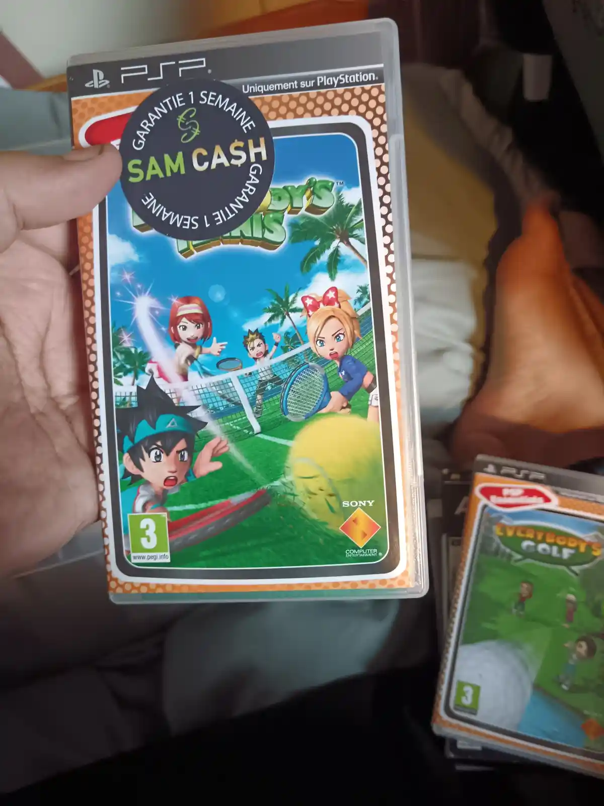A hand holds a PSP game case for 'Everybody's Tennis' featuring cartoon characters on a tennis court. A black circular…