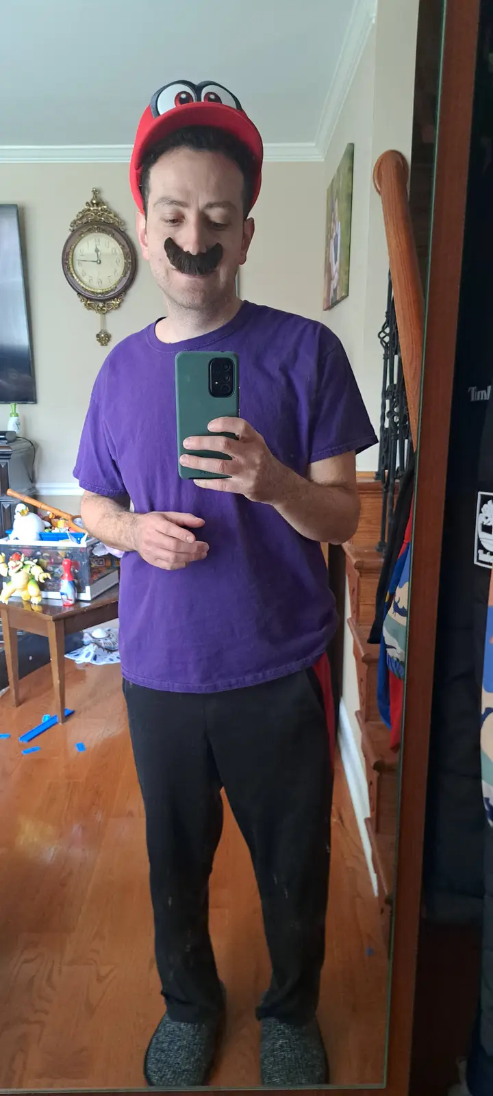A man takes a mirror selfie wearing a red cap with cartoon eyes, a fake thick mustache, and a purple t-shirt. He holds a…