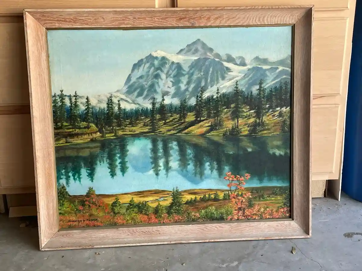 A framed landscape painting by Frances W. Rogers depicts a serene mountain lake scene with snow-capped peaks, evergreen…
