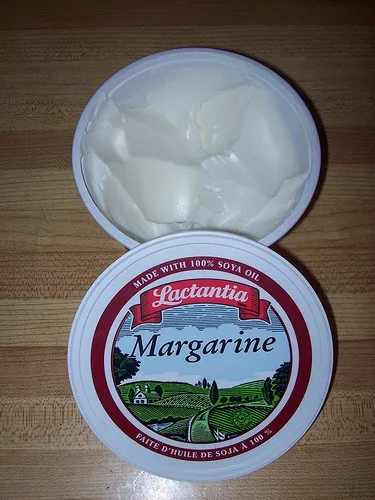 An open tub of Lactantia margarine sits on a wooden surface, revealing its creamy white contents. The lid displays the brand…
