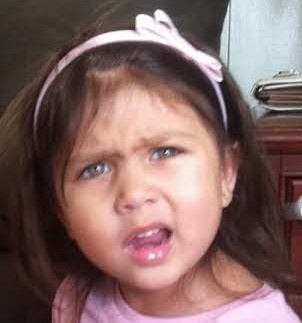 A close-up photo of a young girl with dark hair and a pink headband, making an exaggerated, pouty crying face with her eyes…