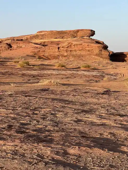 A wide shot of a desert landscape with red sandstone rock formations under a clear blue sky, featuring sparse green shrubs…