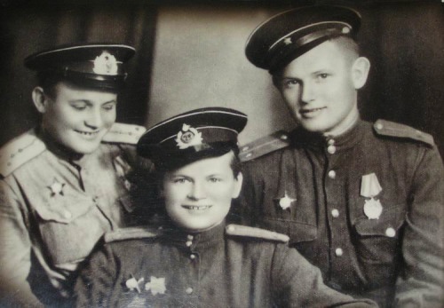 A vintage black-and-white photo shows three smiling young people in Soviet military uniforms, with medals and insignia…