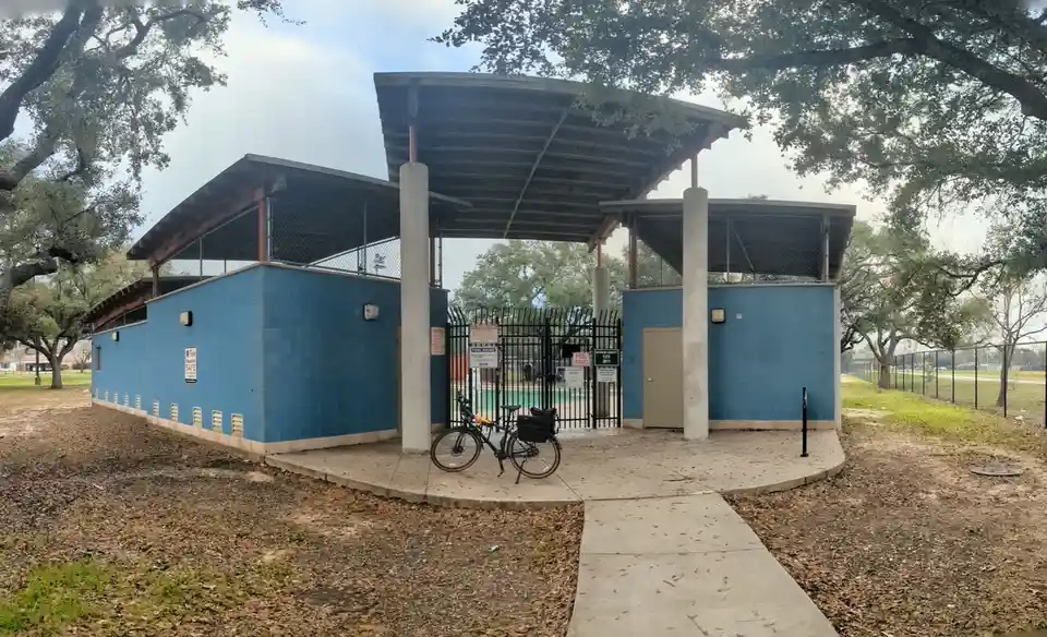 A black bicycle with a rear rack and bag is parked on a concrete pad in front of a blue brick pool entrance building with a…