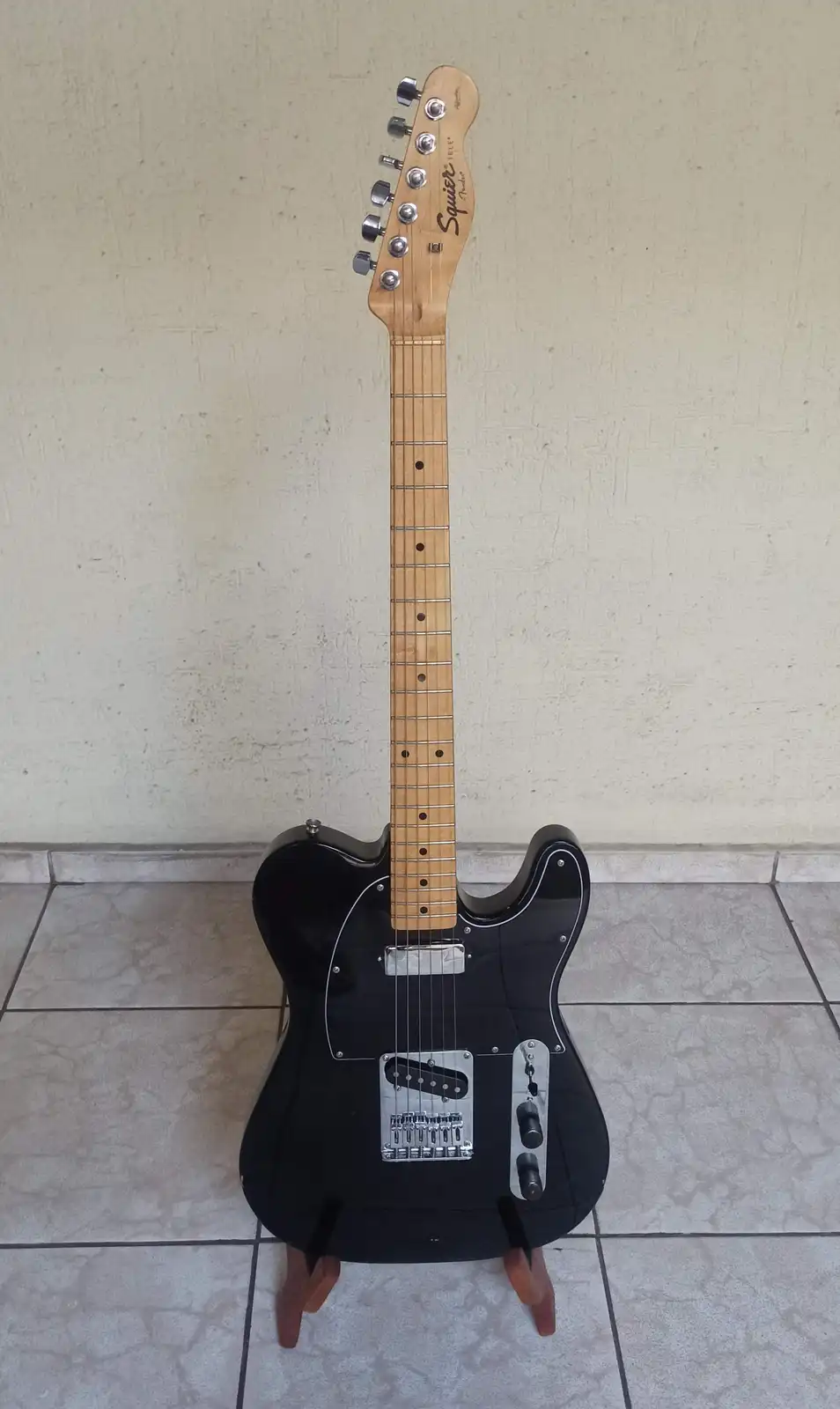 A black Squier Telecaster electric guitar with a natural wood neck and headstock stands upright on a tiled floor against a…