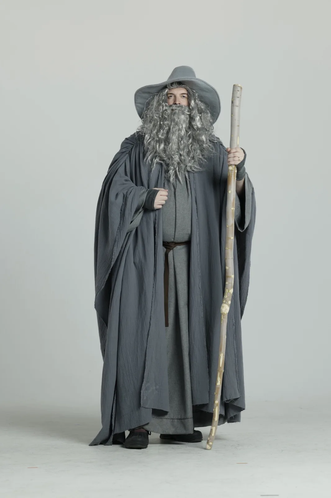 A person dressed as a wizard in a grey robe, wide-brimmed hat, and long grey beard, holding a wooden staff, stands against a…