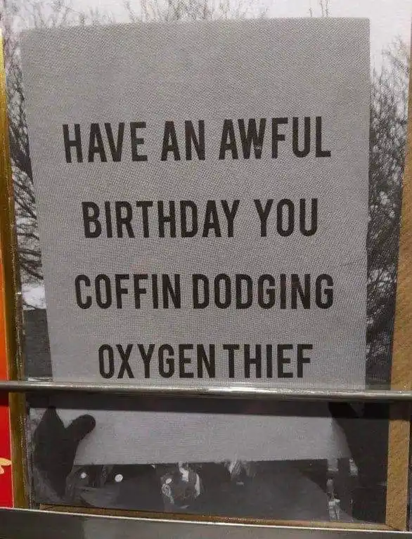 A close-up photo shows a grey sign with bold black text reading 'HAVE AN AWFUL BIRTHDAY YOU COFFIN DODGING OXYGEN THIEF'…