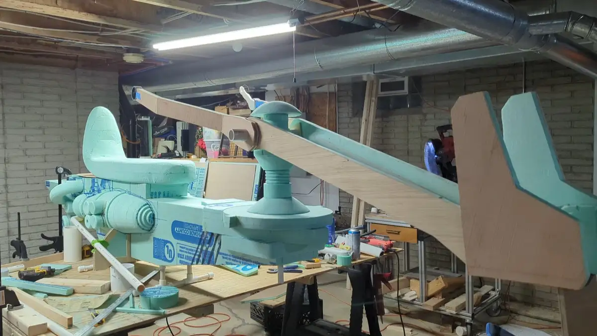 A large, crudely constructed airplane model sits on a workbench in a cluttered garage workshop. The plane is painted light…