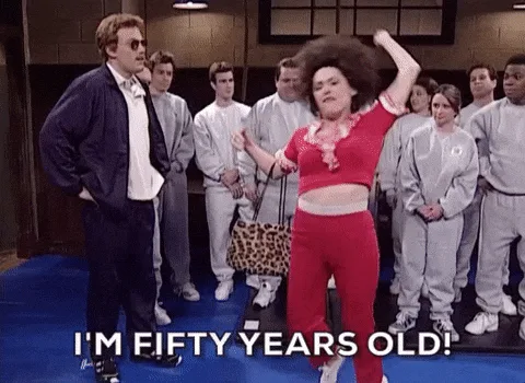 A woman in a red outfit and large afro dances energetically in front of a group of people in grey uniforms, while a man in a…