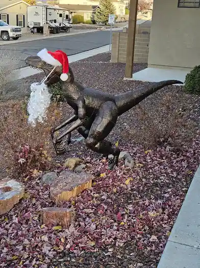 A metal statue of a dinosaur, likely a raptor, is dressed as Santa Claus, wearing a red hat with white trim and holding a…