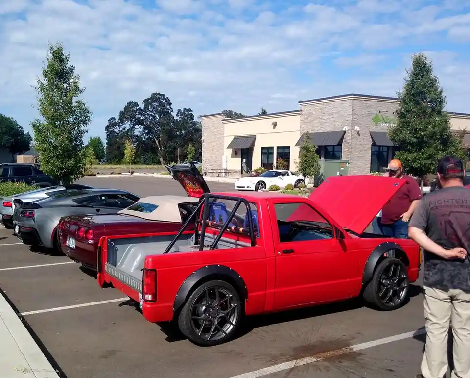 A red pickup truck with its hood open is parked in a lot alongside other cars, including a silver and a maroon sports car…