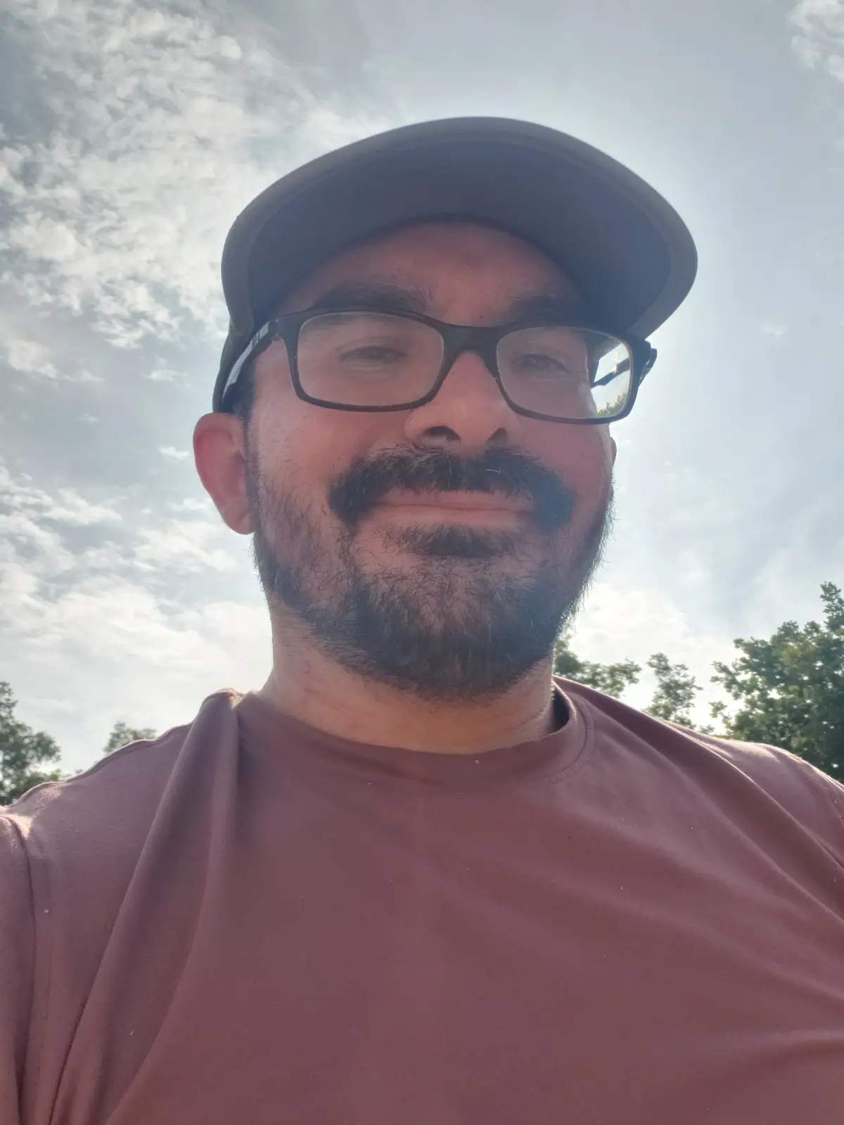 A man with glasses and a cap smiles at the camera against a bright sky with scattered clouds. He has a beard and mustache…