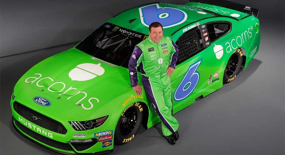 A man in a green racing suit leans against a bright green Ford Mustang race car with the number 6 and 'acorns' branding. The…