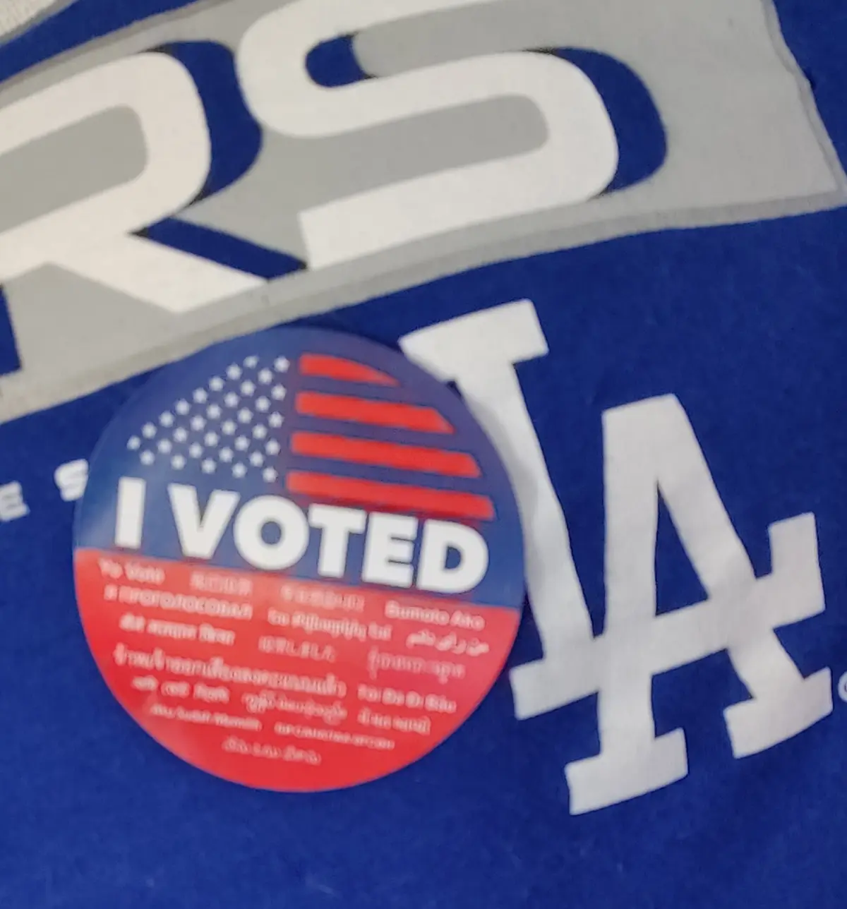 A close-up of a circular 'I VOTED' sticker with an American flag design is stuck on a blue Los Angeles Dodgers t-shirt. The…