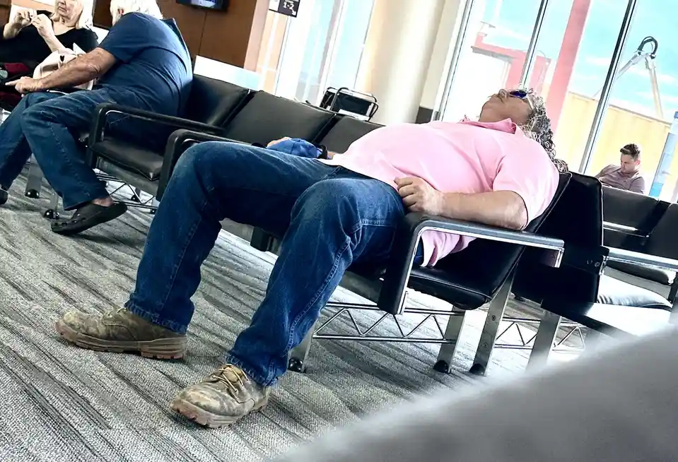 A man in a pink shirt and blue jeans is sleeping on a black airport chair, his head resting on the armrest, wearing dirty…