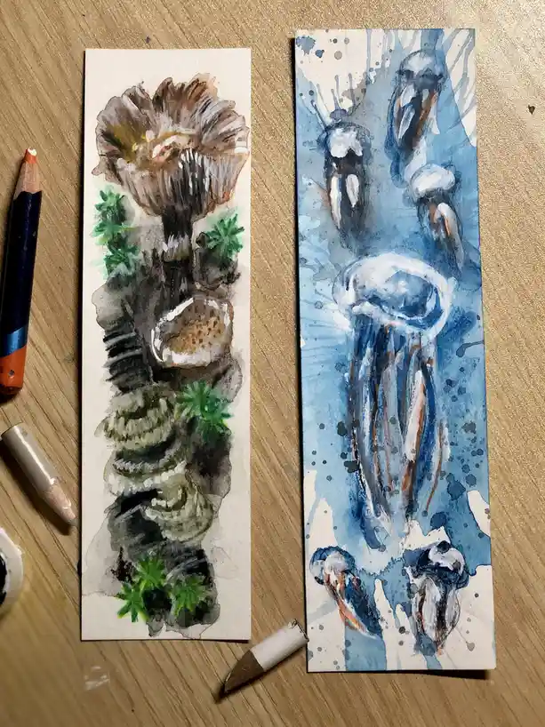 Two watercolor paintings on rectangular paper sit side-by-side on a wooden surface. The left painting depicts a brown…