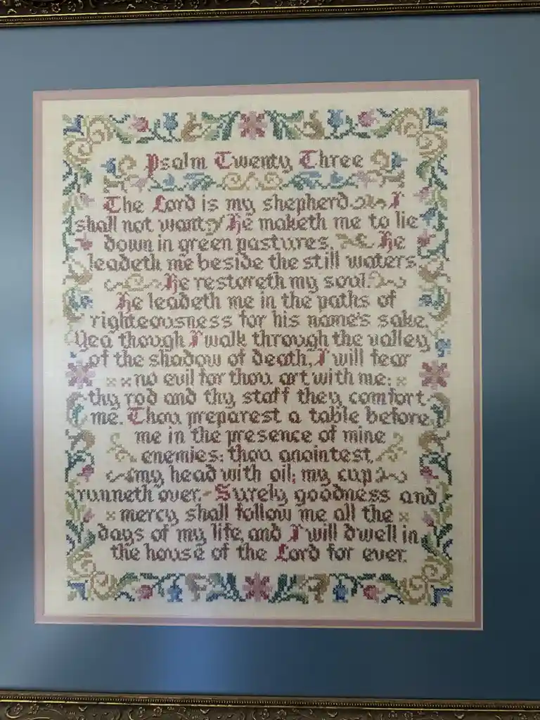 A framed, colorful cross-stitch artwork displays the full text of Psalm 23, with decorative floral borders, mounted on a…