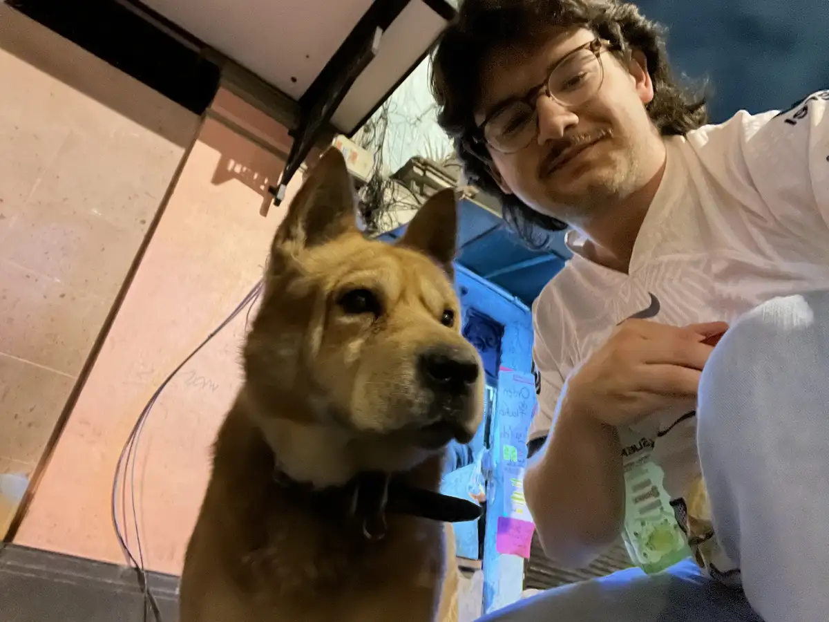A low-angle selfie shows a person with glasses and a white shirt sitting beside a tan dog wearing a black collar. The dog…