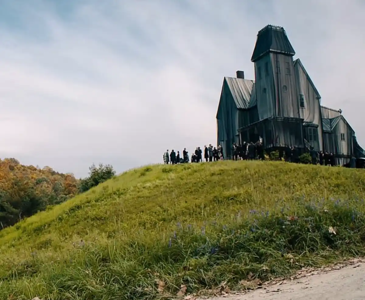 A group of people stands on a grassy hill in front of a large, dilapidated, and oddly shaped house with a dark, weathered…