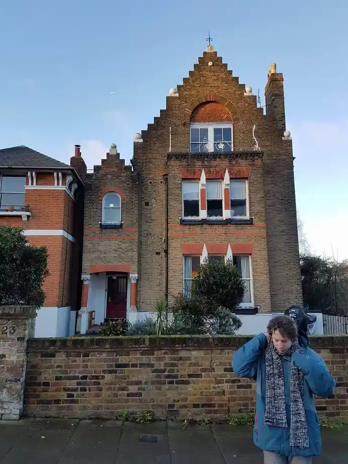 A person in a blue winter coat and scarf stands in front of a large, ornate brick house, adjusting their scarf. The house…