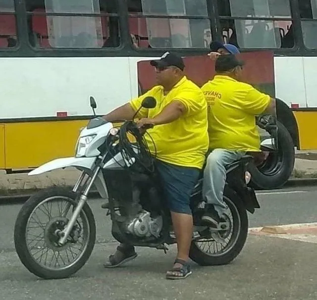 Two men in matching yellow shirts ride a motorcycle; the passenger's shirt reads 'CHAVE' on the back. A bus is visible…