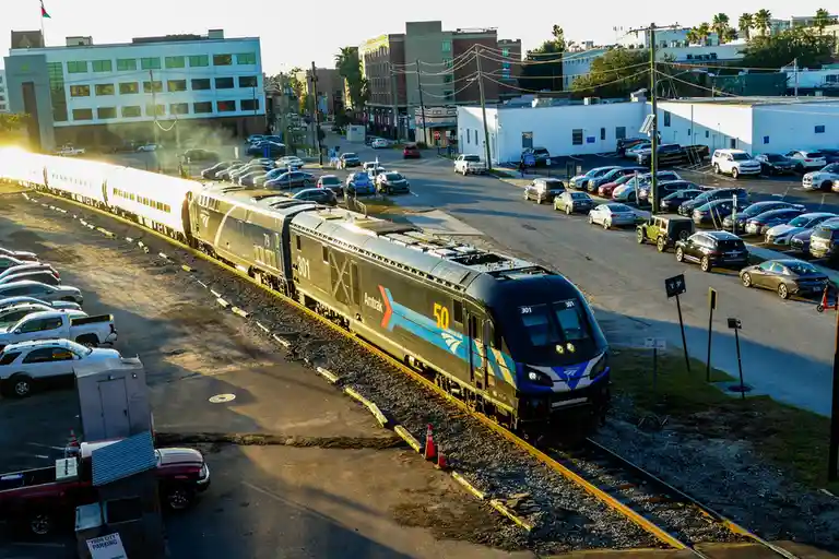 An Amtrak train, numbered 301, travels on tracks adjacent to a large parking lot filled with numerous parked cars. The scene…