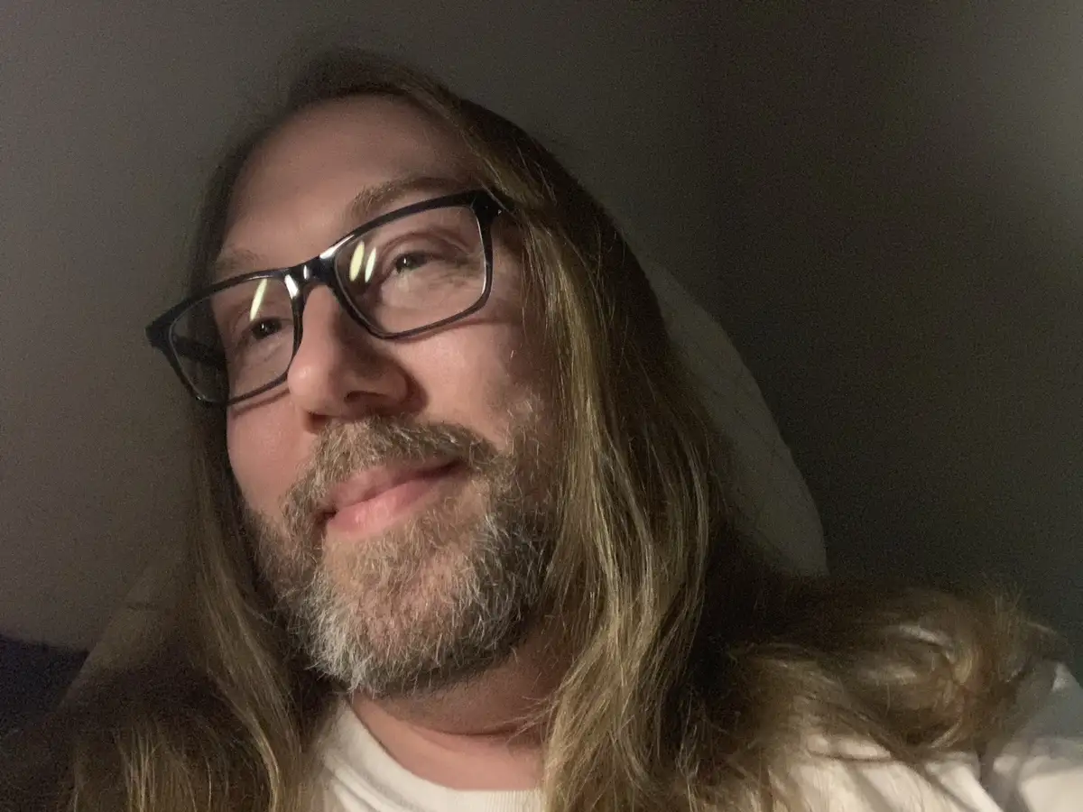 A close-up selfie of a man with long blonde hair, a graying beard, and black-rimmed glasses, looking slightly upward with a…