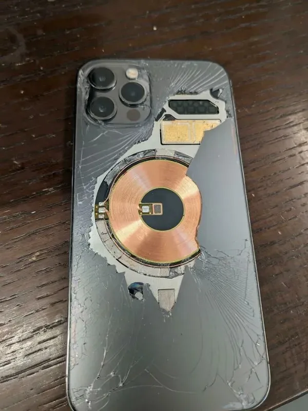 A severely damaged smartphone lies on a wooden surface, its back glass shattered and cracked in a spiderweb pattern. The…