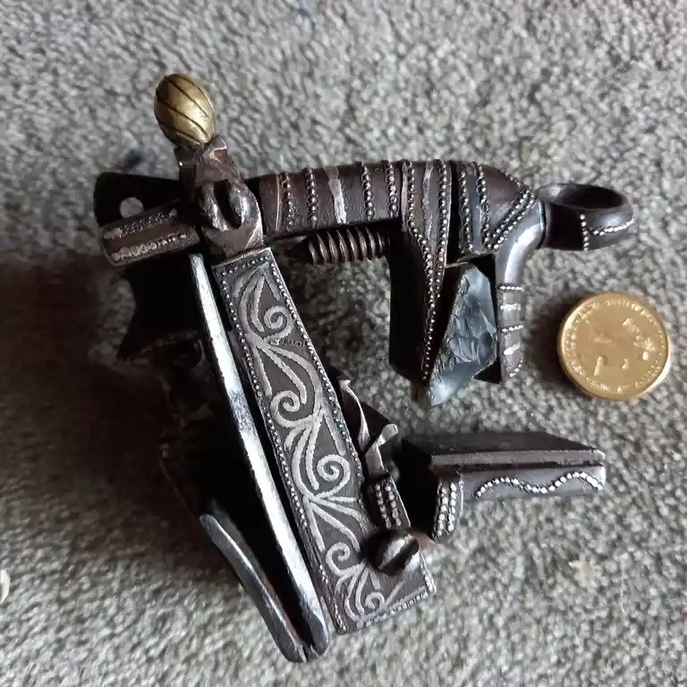 A small, ornate, antique-style pistol with intricate silver scrollwork and a brass knob on top lies on a textured gray…