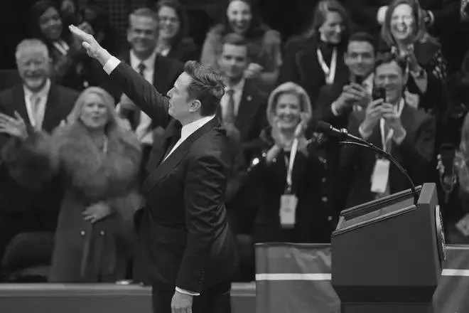 A black-and-white photo shows a man in a suit waving to an audience while standing at a podium. The crowd behind him is…
