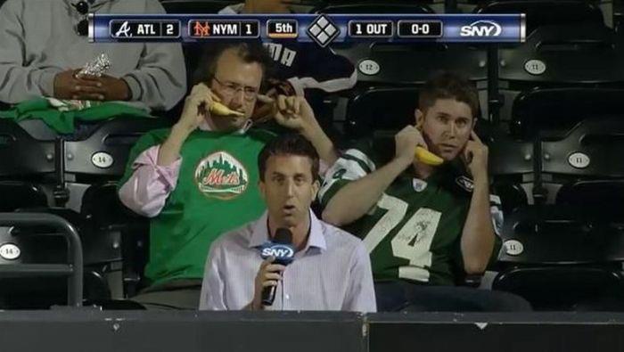 two men in green sports jerseys are sitting in stadium seats, each holding a banana to their ears as if it were a phone. A…