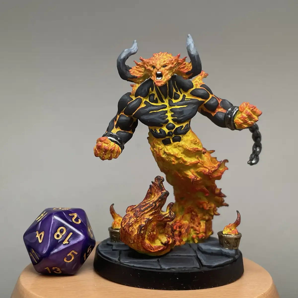 A detailed miniature figurine of a fiery, muscular demon with black horns and a roaring expression stands on a stone base…