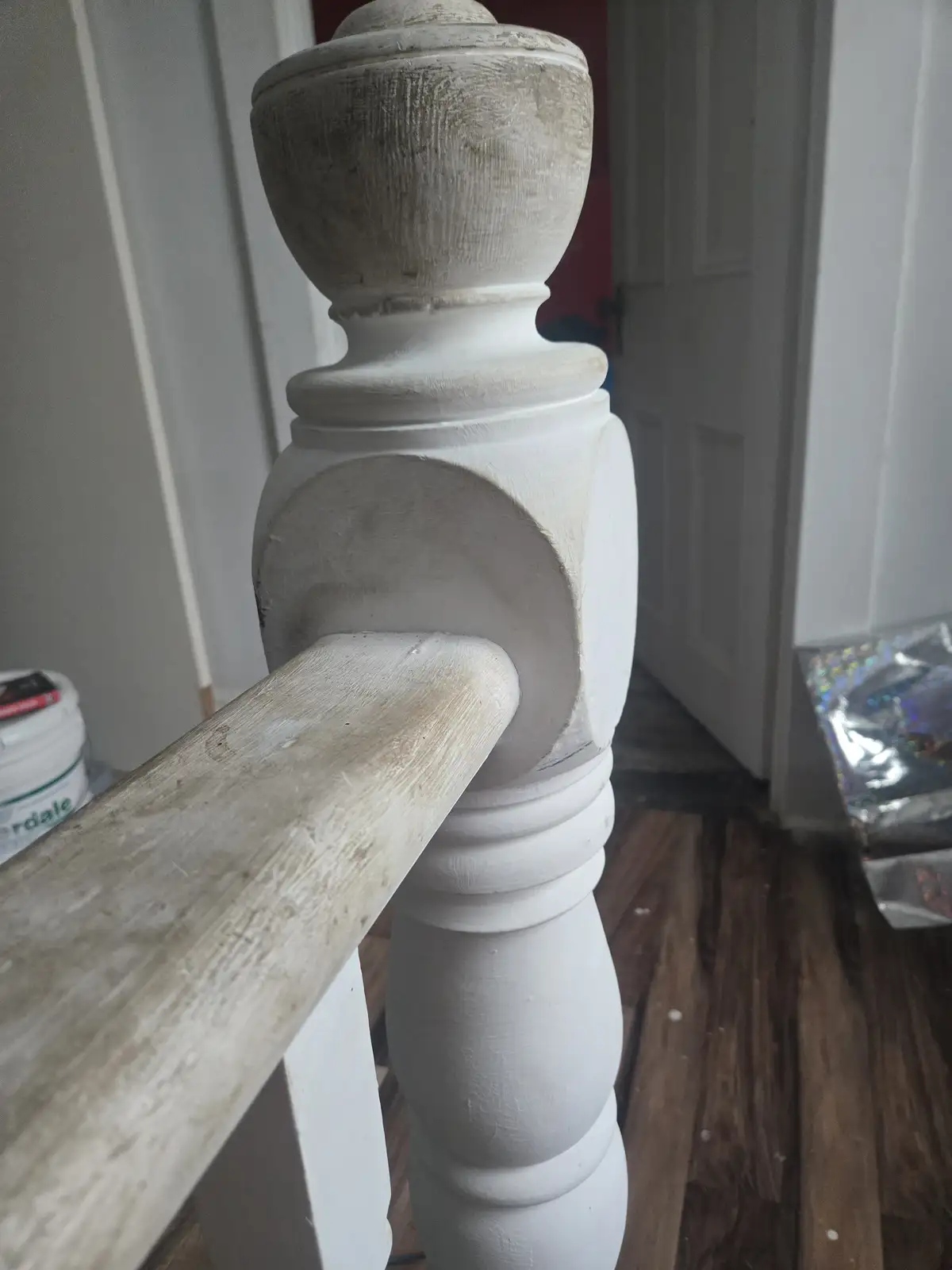 A close-up photo shows a freshly painted white wooden staircase banister post and handrail, with visible brush strokes and…