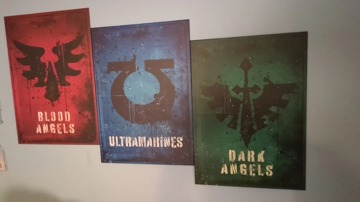Three framed posters hang on a wall, each displaying a different faction's logo and name from the Warhammer 40,000 universe…