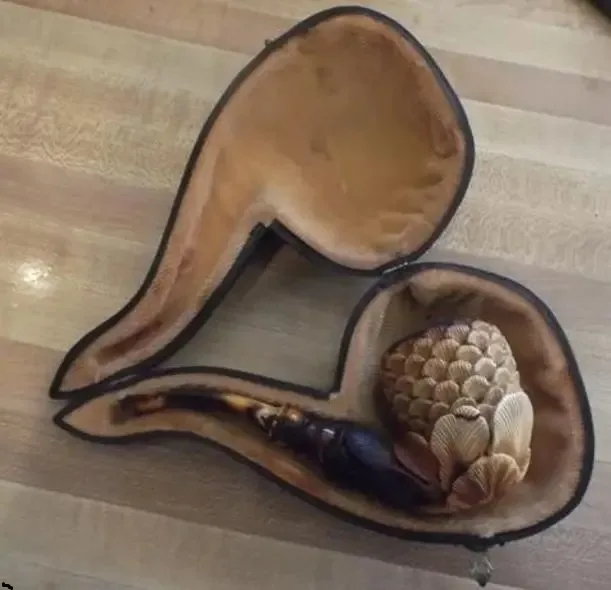 An open, heart-shaped pipe case with a carved wooden pipe inside, resting on a wooden floor. The case is lined with brown…