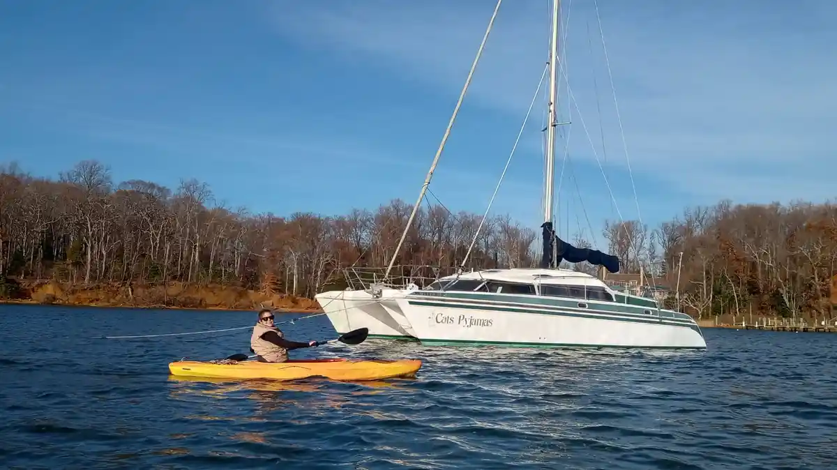 A person in a yellow kayak paddles on calm water near a large white catamaran sailboat named 'Cats Pyjamas'. The boat has a…