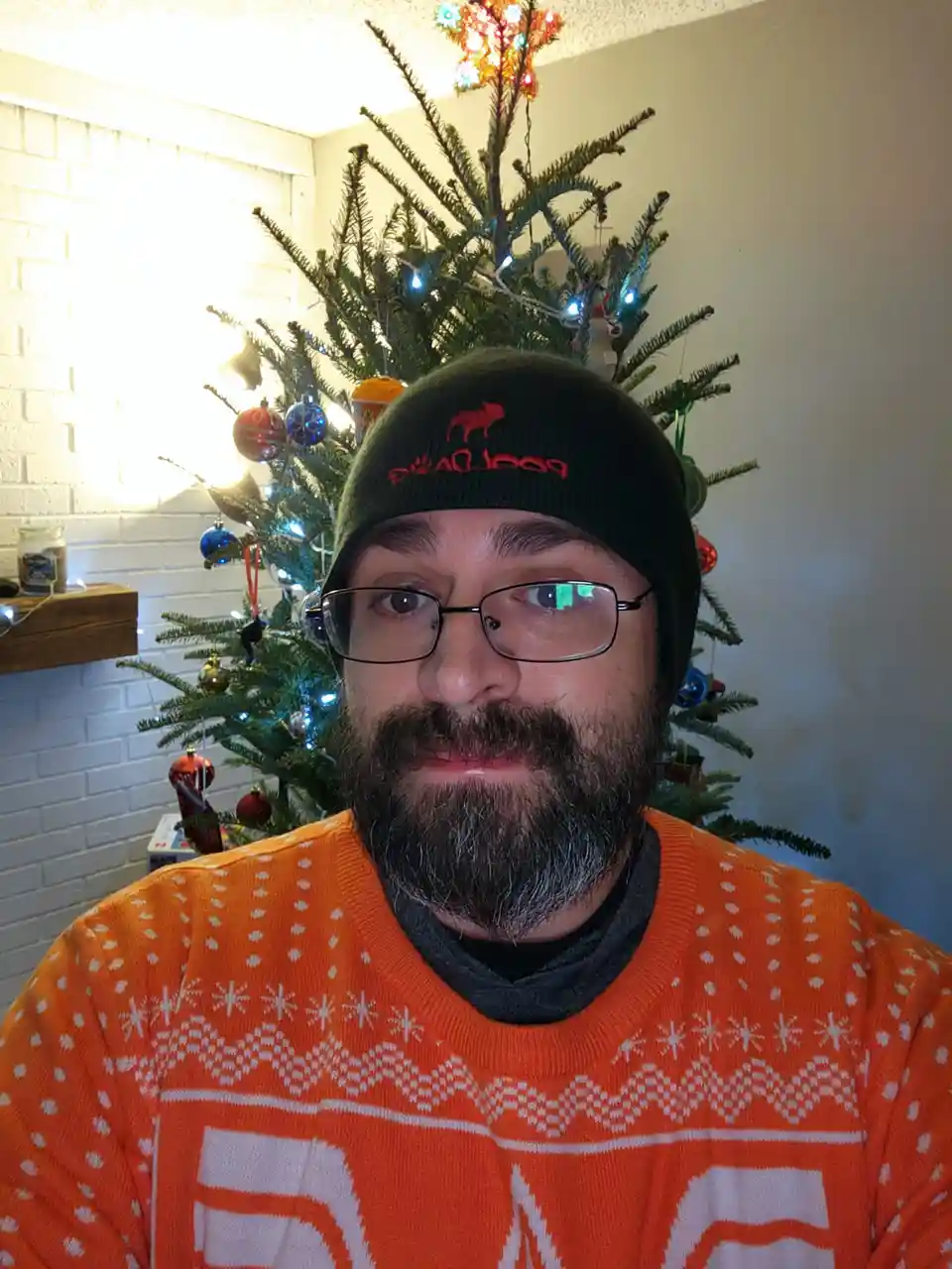 A man with a beard and glasses wears a black beanie and an orange holiday sweater while standing in front of a decorated…
