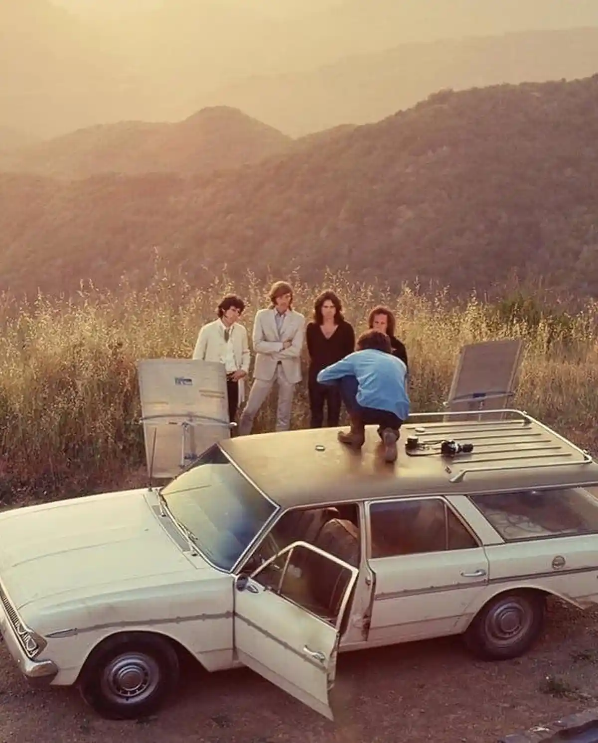 A vintage light-colored station wagon is parked on a dirt road with mountains in the background. One person in a blue shirt…