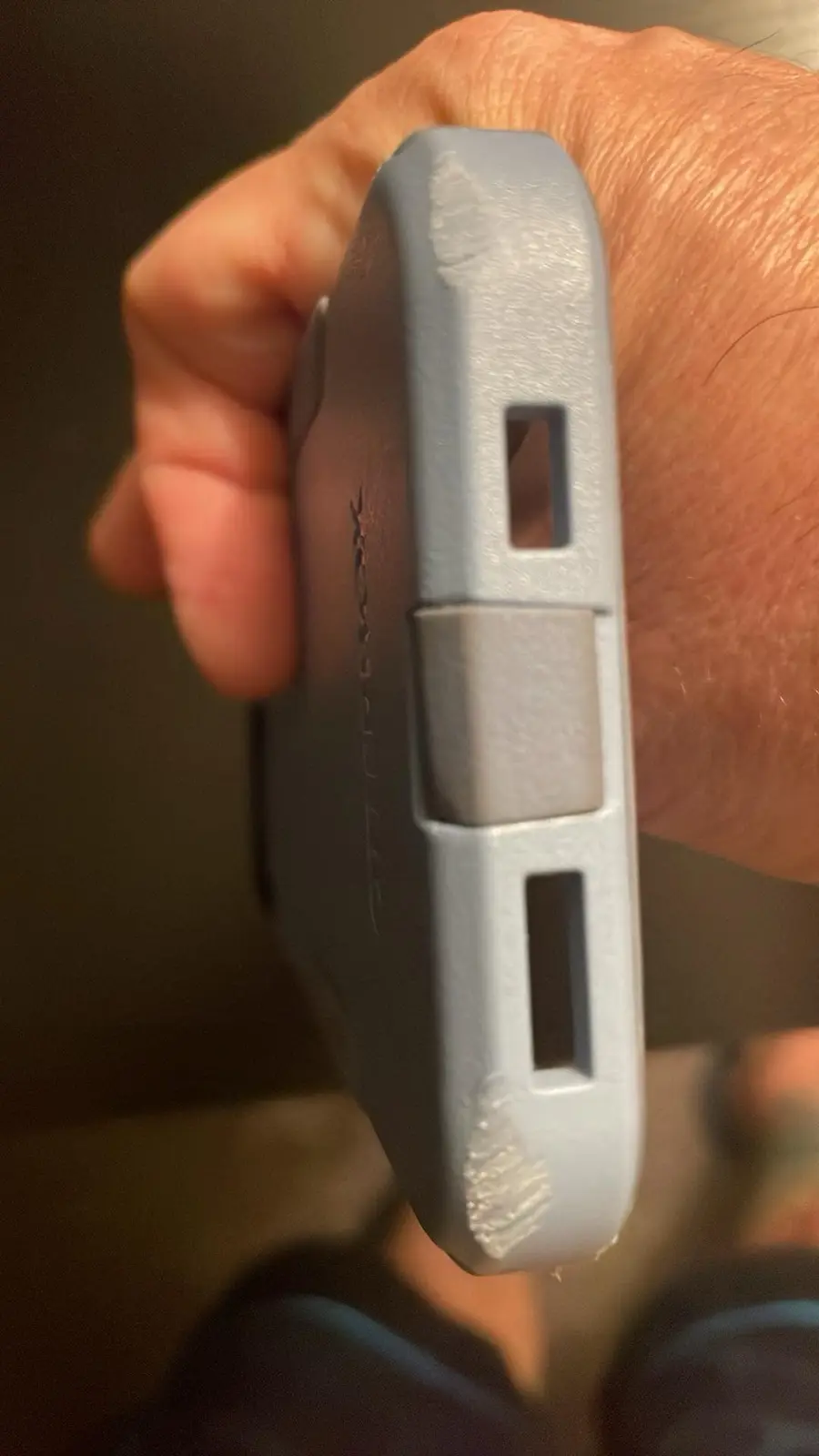 A close-up photo shows a person's hand holding a gray phone case, highlighting its side with two rectangular cutouts and a…